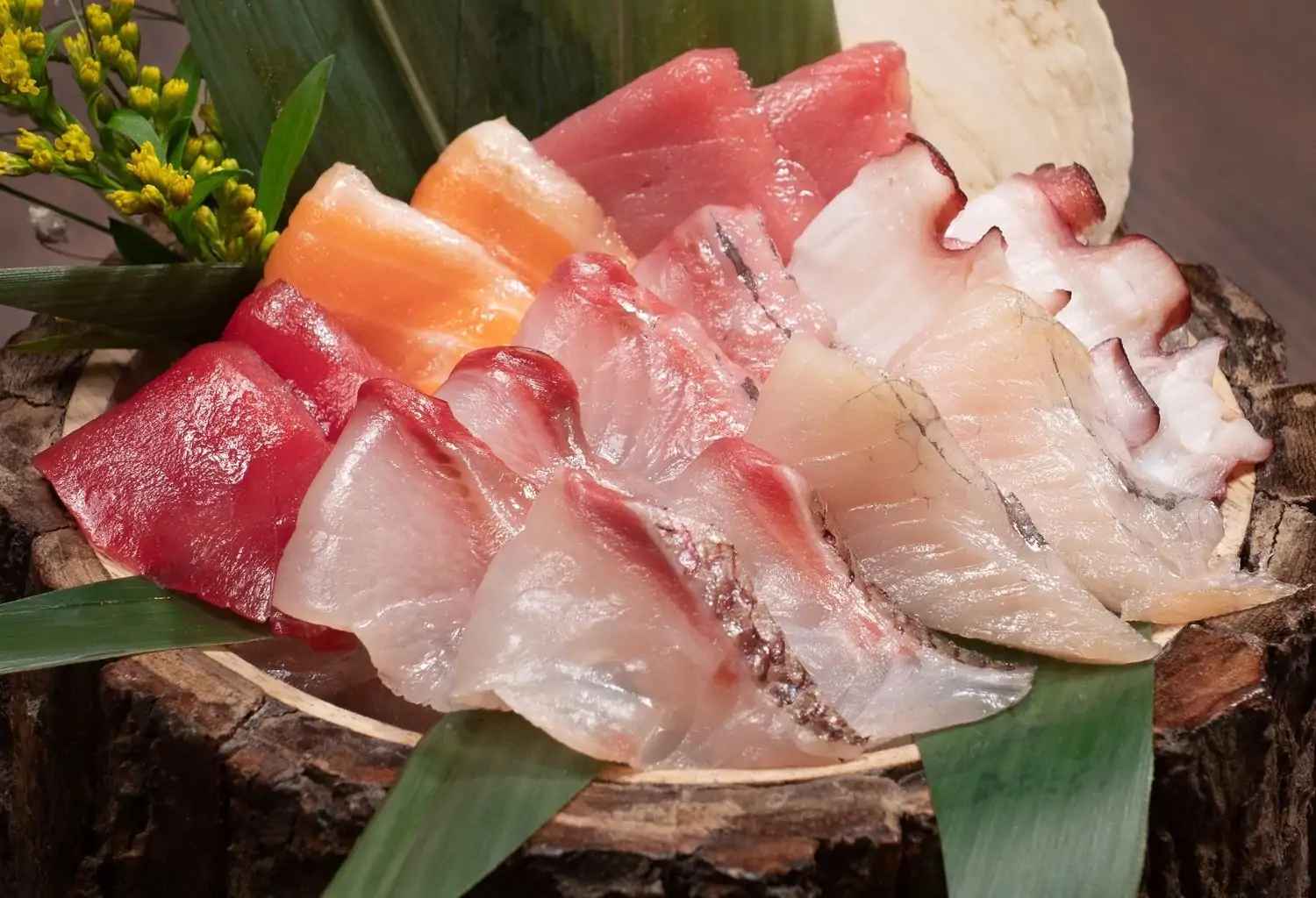 Close-up of a sashimi platter with tuna, salmon, white fish, and octopus, arranged on a rustic wooden platter with green leaves and yellow flowers at one of OpenTable’s most popular Japanese restaurants in Chicago, Juno.