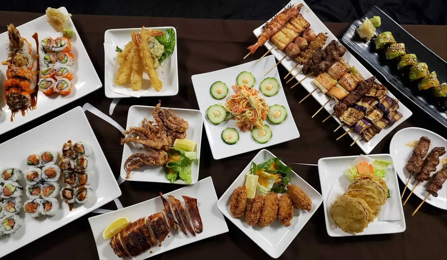 A variety of Japanese dishes on a brown table, featuring sushi rolls, tempura shrimp and vegetables, grilled meat and vegetable skewers, fried squid, grilled fish, and other small plates at Yakitori Boy Japas, one of the best Japanese restaurants in Philadelphia you can book on OpenTable.