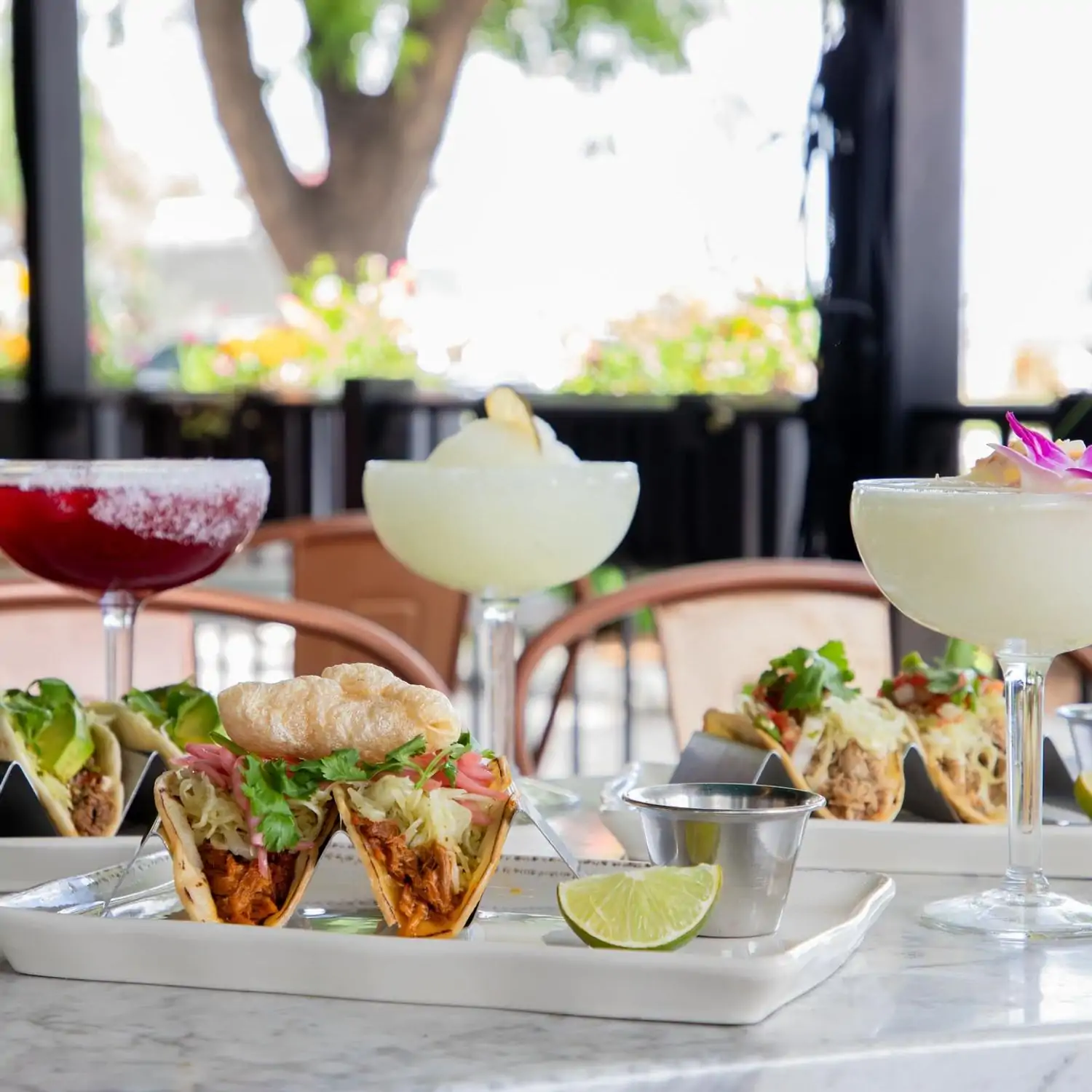 A picture of three large margaritas alongside beautiful platters of tacos accompanied by a lime wedge and sauce in a small silver condiment dish at My Neighbor Felix (LoHi), Denver. Book on OpenTable.