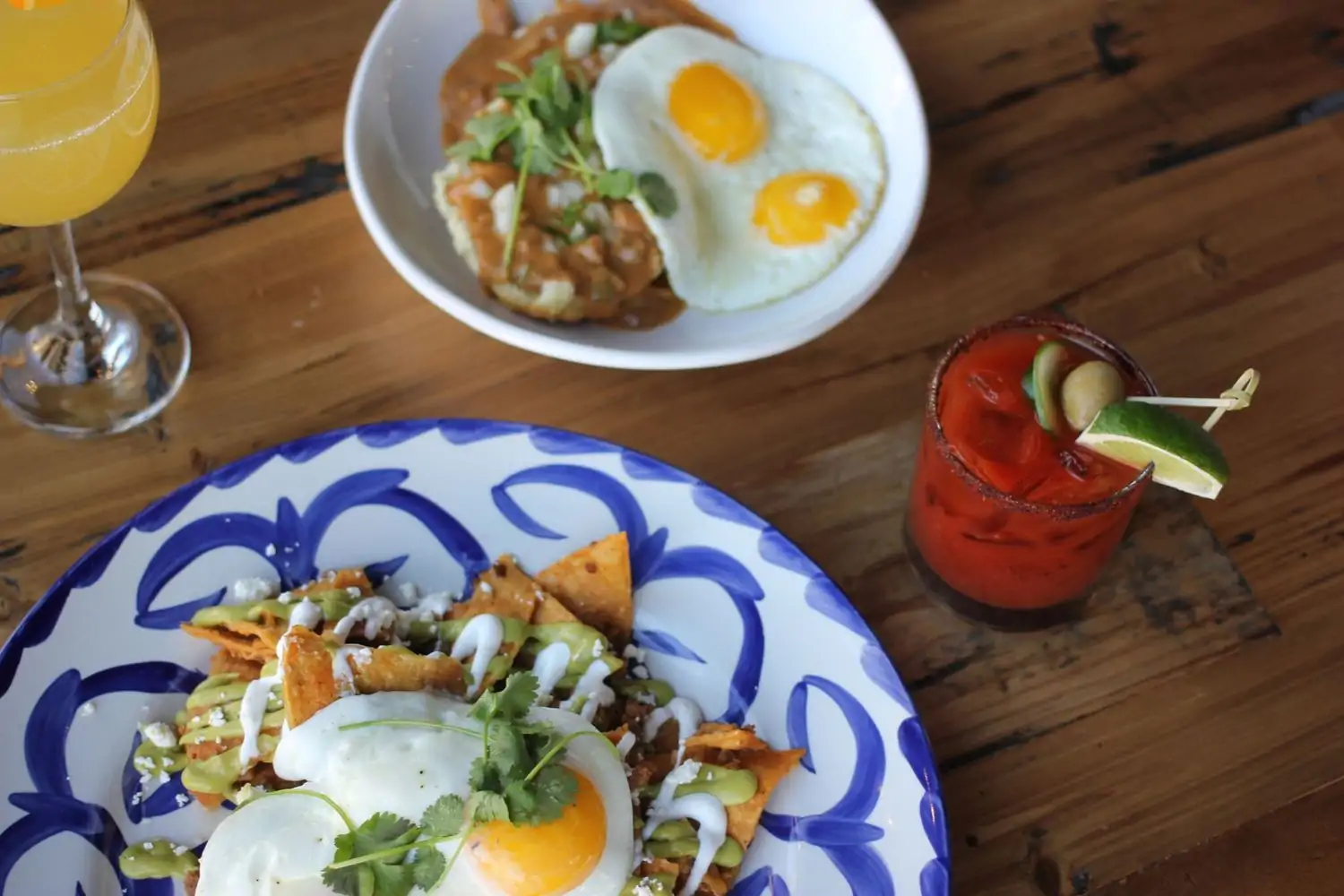 A table has two plates on it, one plate has eggs with mole sauce, and the other has crispy tortilla chips topped with a sunny side up egg, green sauce, white sauce, and fresh cheese, two colorful drinks are also present at El Jefe (Highlands), one of the best Mexican restaurants in Denver. Book on OpenTable.