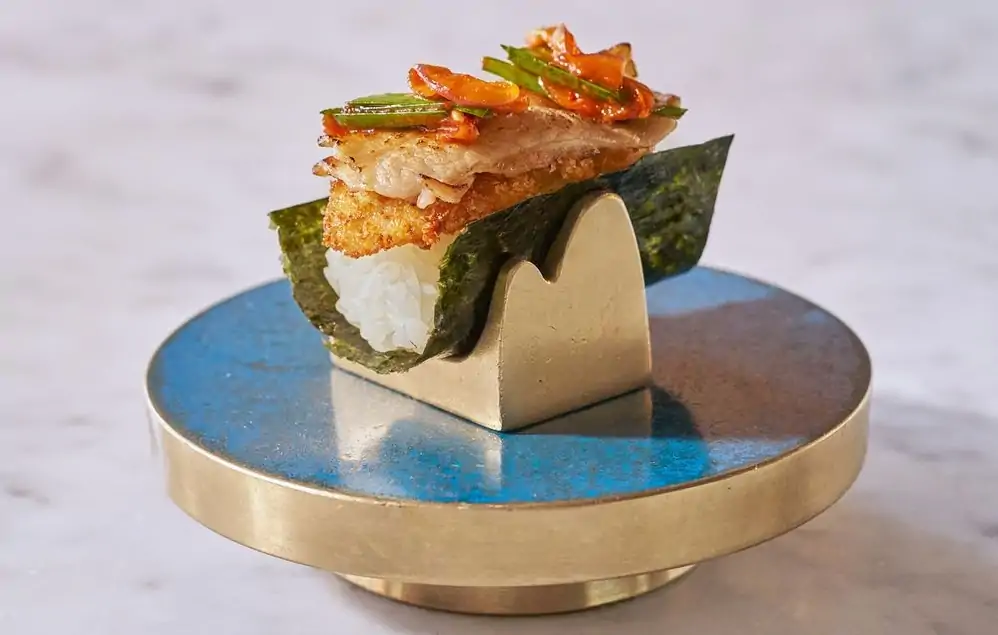 An extreme closeup shows an open hand roll of fried oyster with red and green vegetable topping resting in a bronze holder on a blue enamel disc with a bronze rim at Mari, NYC, available on OpenTable.