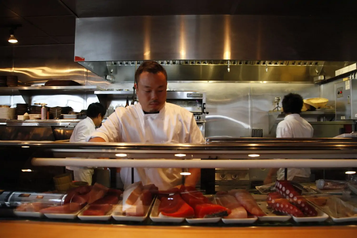 A sushi chef, clad in a crisp white coat, meticulously prepares fresh seafood behind a sleek glass display filled with vibrant cuts of fish and octopus. The open kitchen shows stainless steel surfaces at Sushi Kappo Tamura - Dining Room. Book the best Japanese restaurant in Seattle on OpenTable.