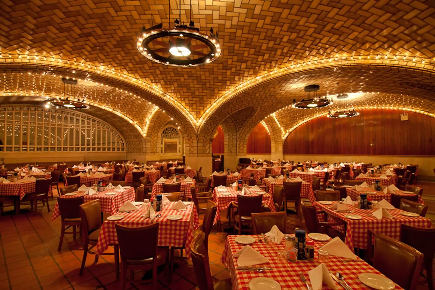 A sea of square dining tables with red and white checked tablecloths fills a room under vaulted barrel arches of patterned tile and lined with strings of Edison bulbs, giving the room an amber glow at Grand Central Oyster Bar, an OpenTable restaurant for some of the best solo dining in NYC.