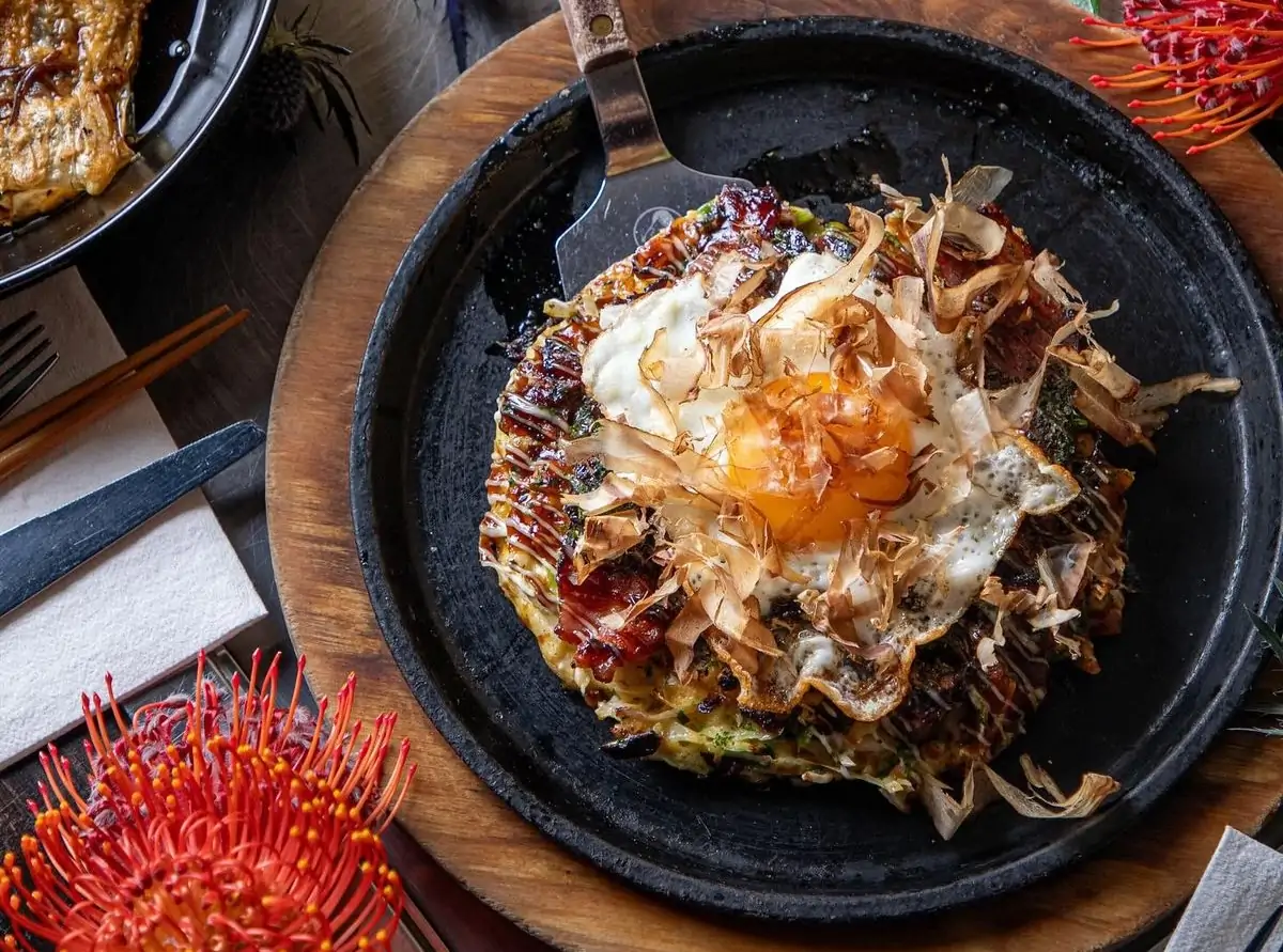 A savory Japanese pancake (okonomiyaki) with a fried egg, delicate bonito flakes, and a dark, glossy sauce is presented on a round black cast-iron plate at Chicago's Gaijin—available to book on OpenTable.