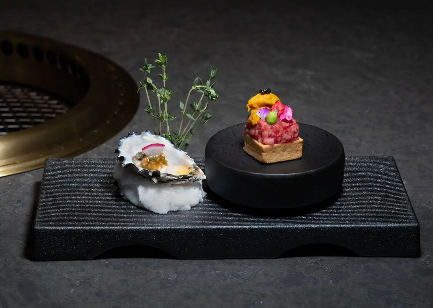 Close-up of a raw oyster on crushed ice with a garnish and a small tartare dish on a round black stand, presented on a rectangular textured black plate at Itadaki Yakiniku, one of the best Japanese restaurants in Seattle, available on OpenTable.