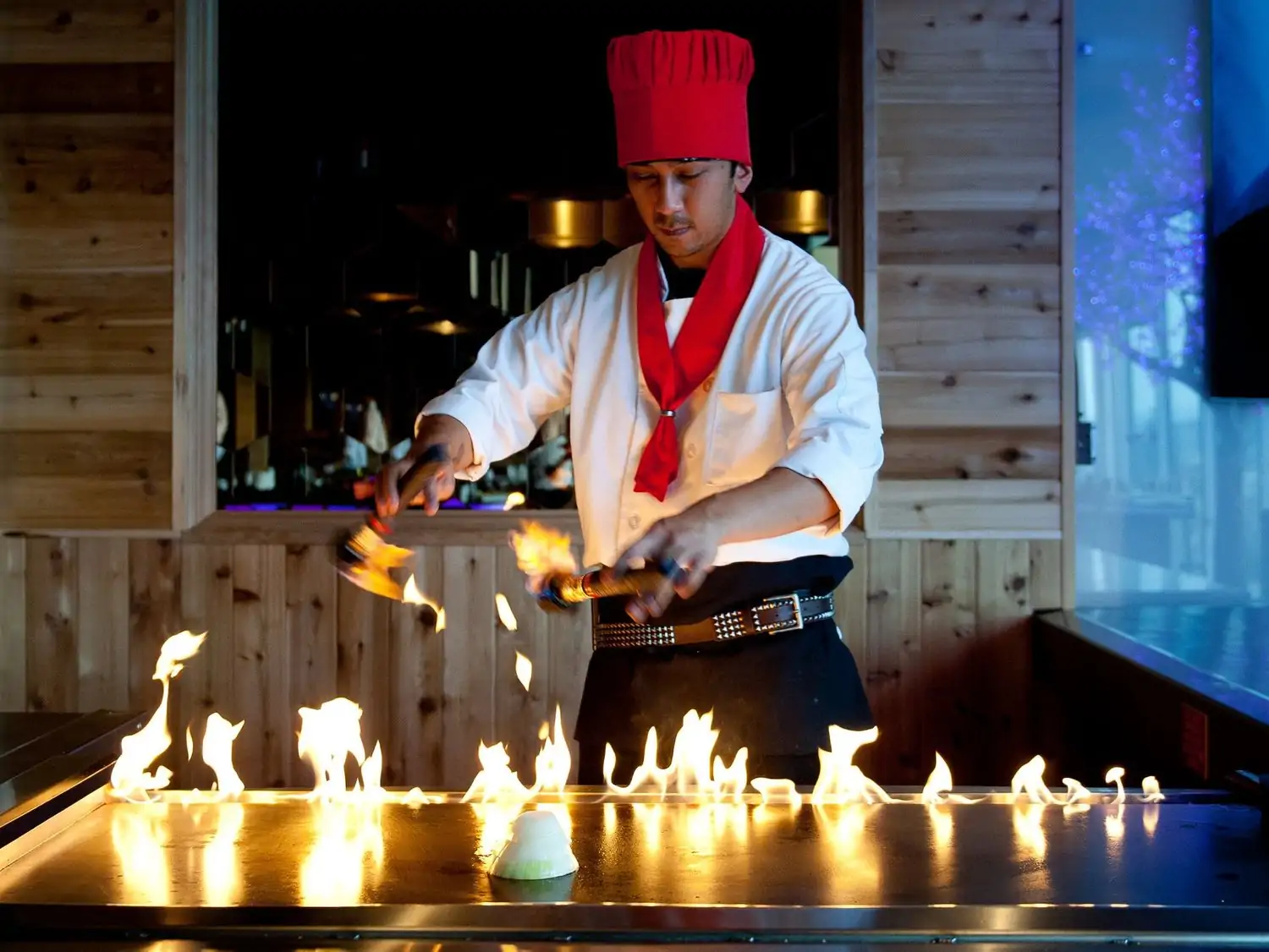 A teppanyaki chef in a red hat and scarf wields cooking utensils over a flaming hot grill. Reflections of the flames dancing across the grill at Wild Wasabi Signature, in Seattle. Book on OpenTable.