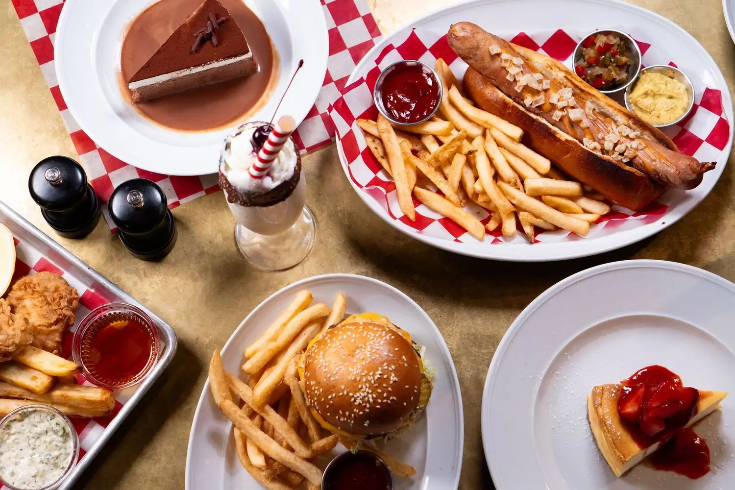 White plates, some with red and white checked paper liners, hold classic diner dishes of hotdogs and hamburgers with French fries, chicken fingers, and cheesecake with a milkshake on a table at B&L Diner, an OpenTable restaurant for the best solo dining in Midtown NYC.