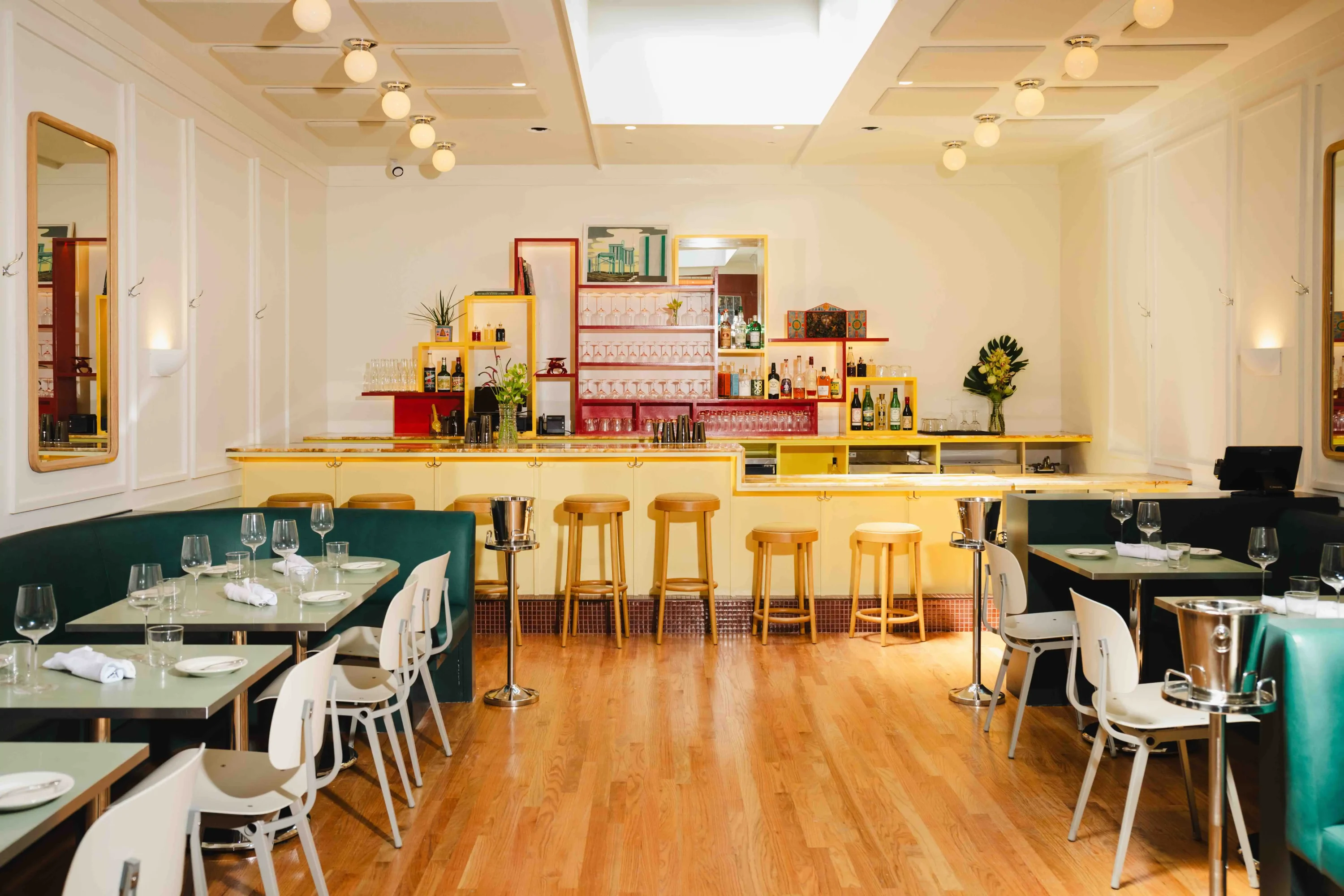 A white-walled dining room with steel tone tables and leather banquettes of various colors sit in front of a pale yellow bar in front of red and yellow shelves holding various bottles and glasses at Strange Delight, an OpenTable oyster bar among the best places for solo dining in NYC.