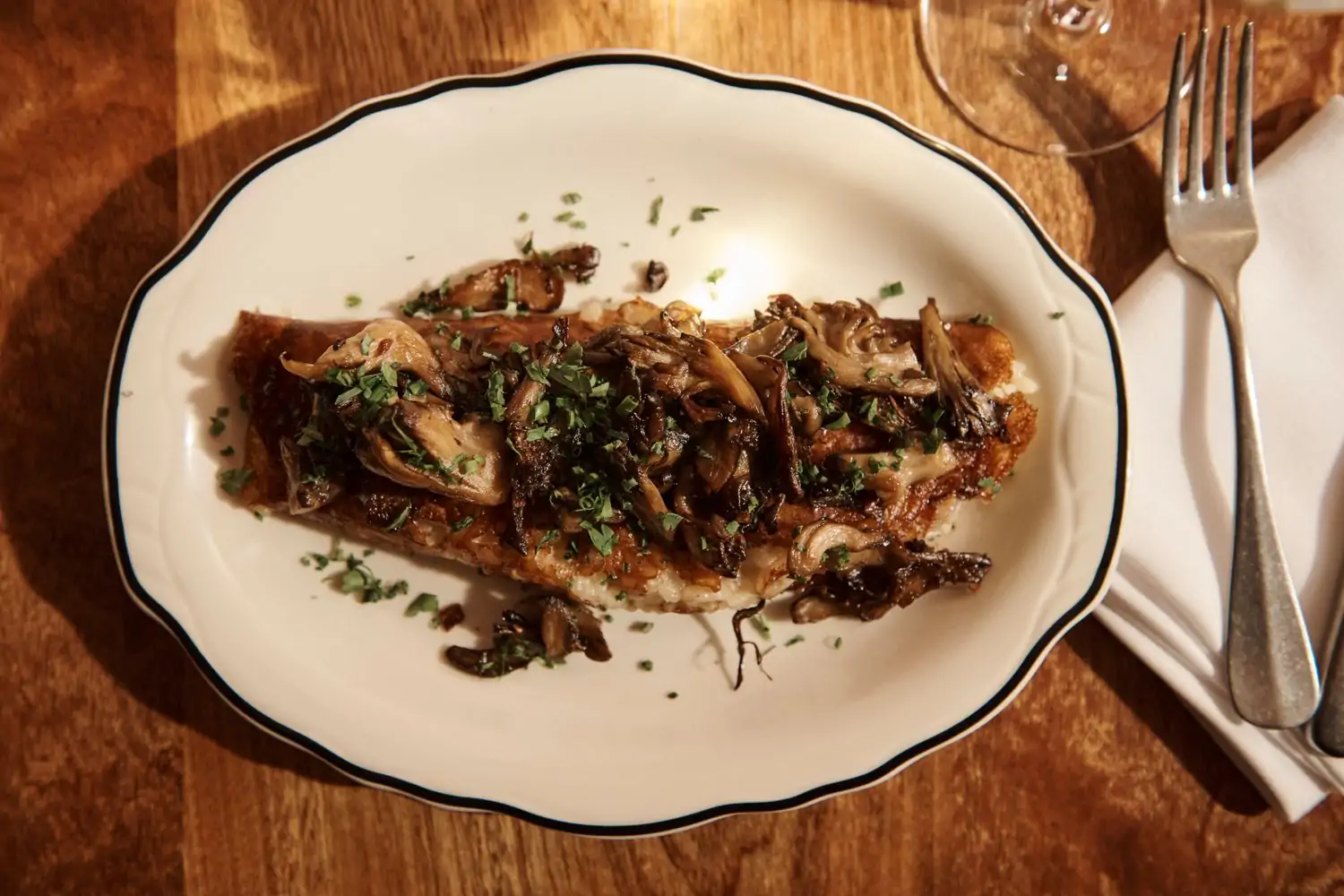 An oval plate holds browned risotto rice topped with king oyster mushrooms on a wooden table with a fork and cloth napkin at Demo, an OpenTable wine bar with some of the best solo dining in NYC.