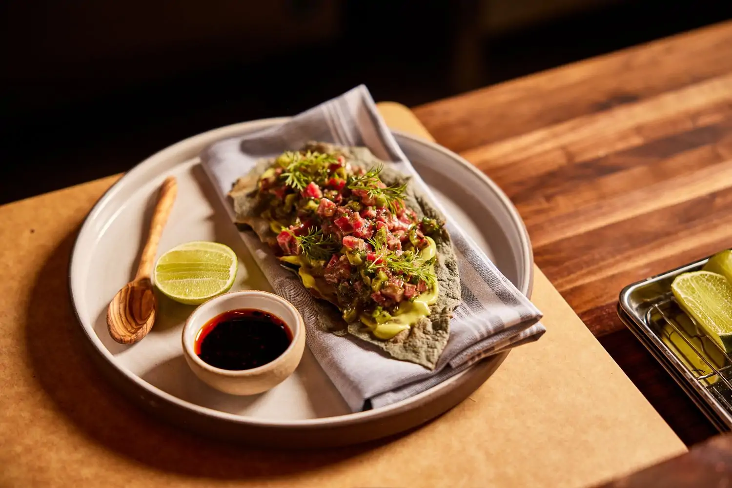 A ceramic plate has a lime wedge, a small wooden spoon, a side of salsa, and an open-faced tortilla is topped with queso sauce, chopped tuna, and fixings at Mezcaleria Alma (LoHi), Denver. Book on OpenTable.