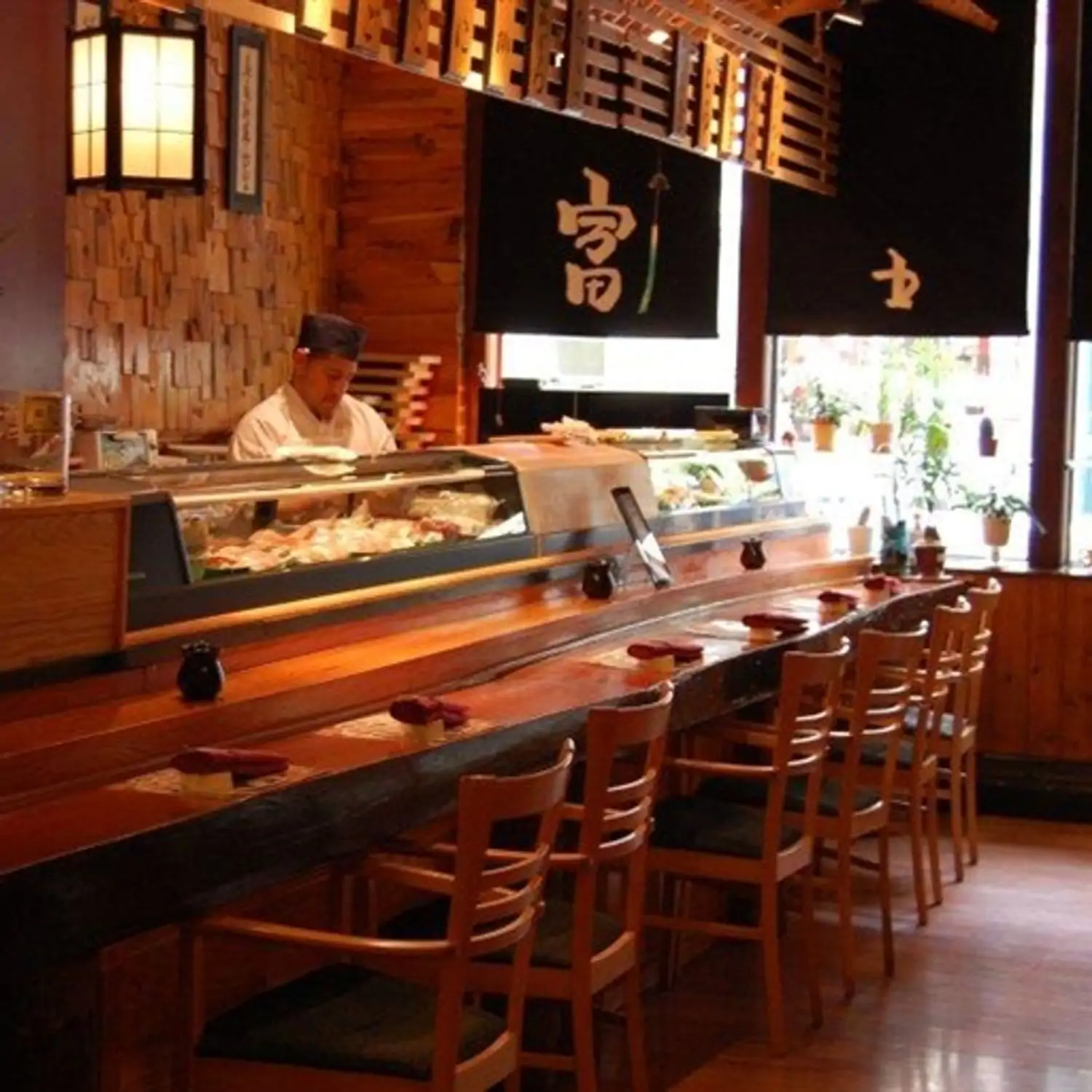 A warm, inviting sushi bar with wooden interiors featuring a long counter where a chef prepares fresh sashimi and sushi behind a glass display filled with seafood. Traditional Japanese decor, soft lighting, and neatly arranged seating at Fuji Mountain Japanese Restaurant, one of the restaurants in Philadelphia you can book on OpenTable.