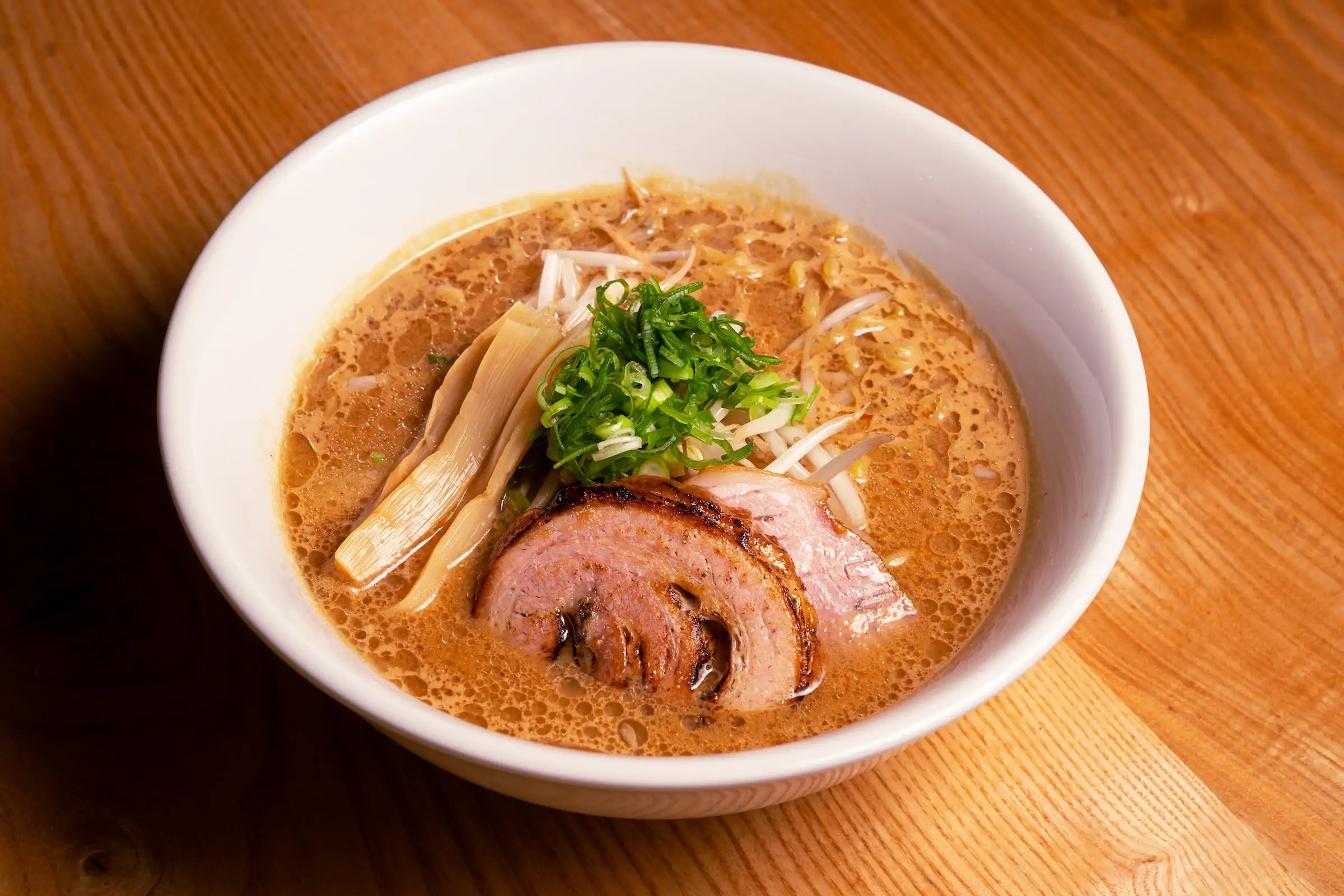 A bowl of ramen with sliced pork belly (chashu), bamboo shoots, and chopped scallions in a rich, opaque broth, served in a white bowl on a wooden table at Akahoshi Ramen, one of the best Japanese restaurants in Chicago that you can book on OpenTable.