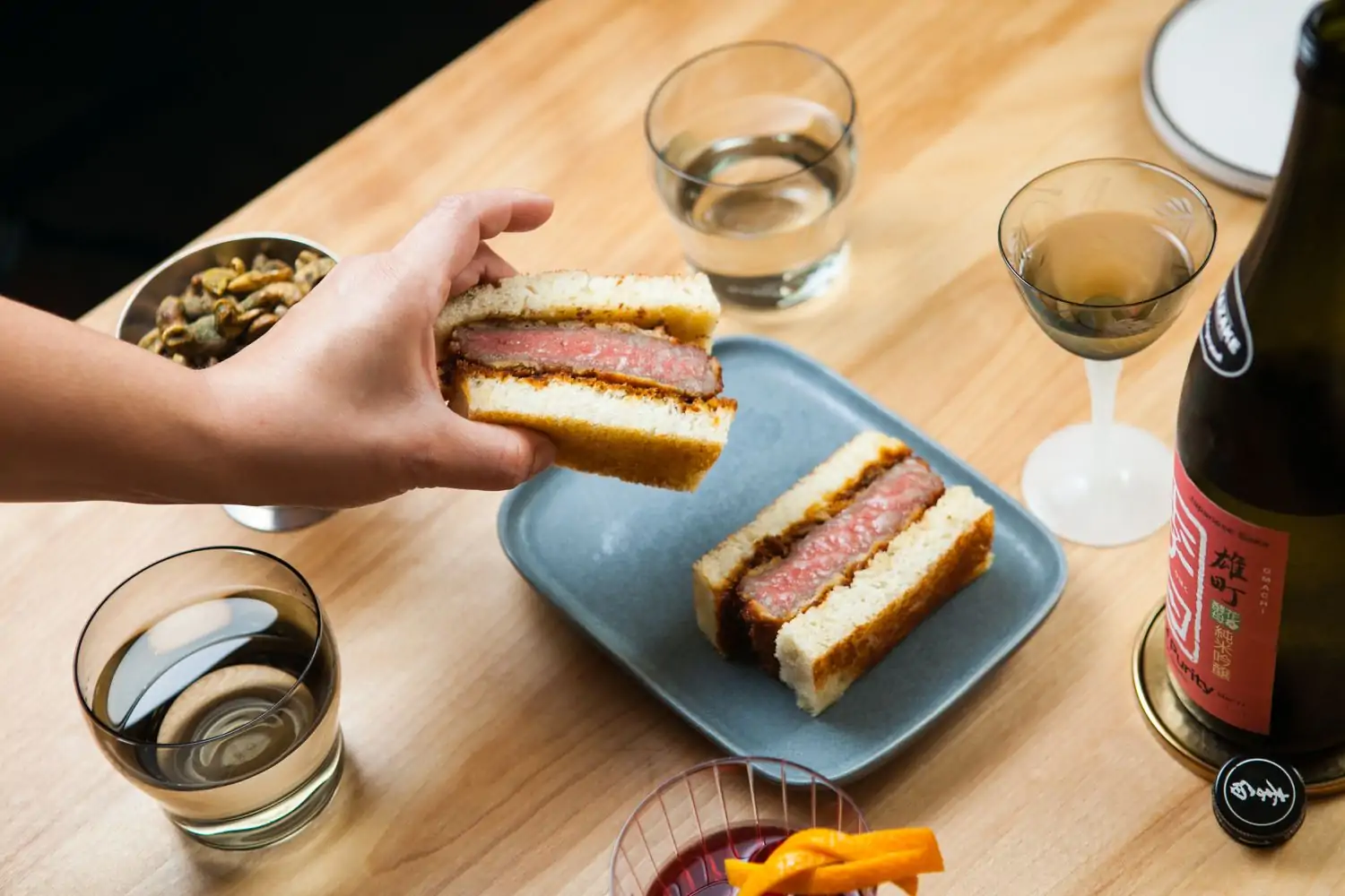 A wagyu katsu sando cut into two pieces on a gray plate, with a bottle of sake, two glasses of water, and a small bowl of snacks, being held by a hand at Kumiko, one of the best Japanese restaurants in Chicago to book on OpenTable.