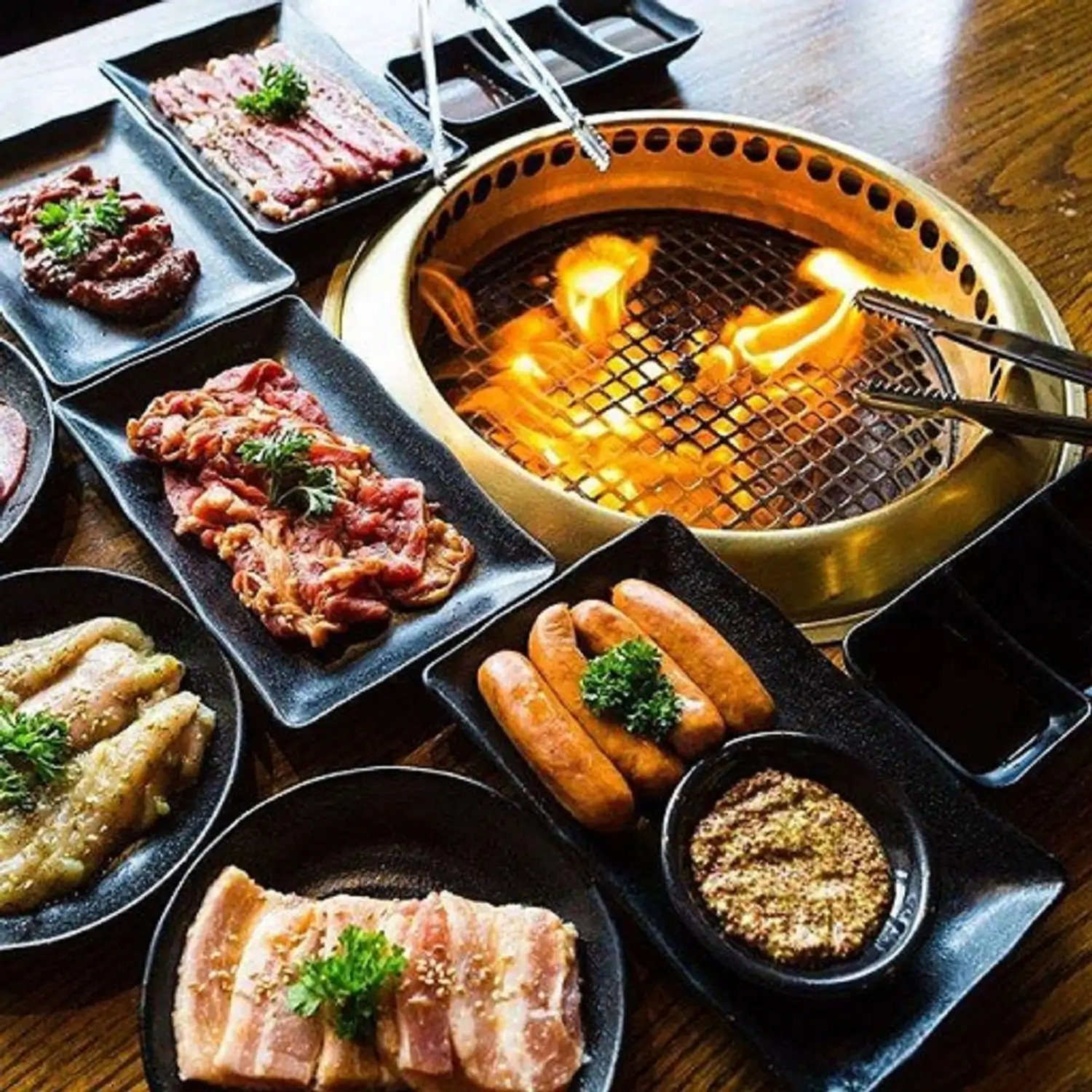 A selection of raw meats, including sliced beef, pork belly, and sausages, artfully arranged on black rectangular plates, surrounding a hot, round grill with visible flames at Gyu-Kaku Japanese BBQ, one of the best Japanese restaurants in Philadelphia—and available to book on OpenTable.