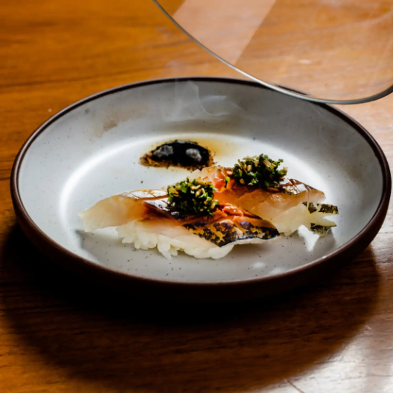 Two pieces of nigiri sushi with seared fish and a green herb garnish, presented on a round, speckled gray plate with a partially visible glass lid at Momotaro, the best Japanese restaurant in Chicago on OpenTable for upscale dining.