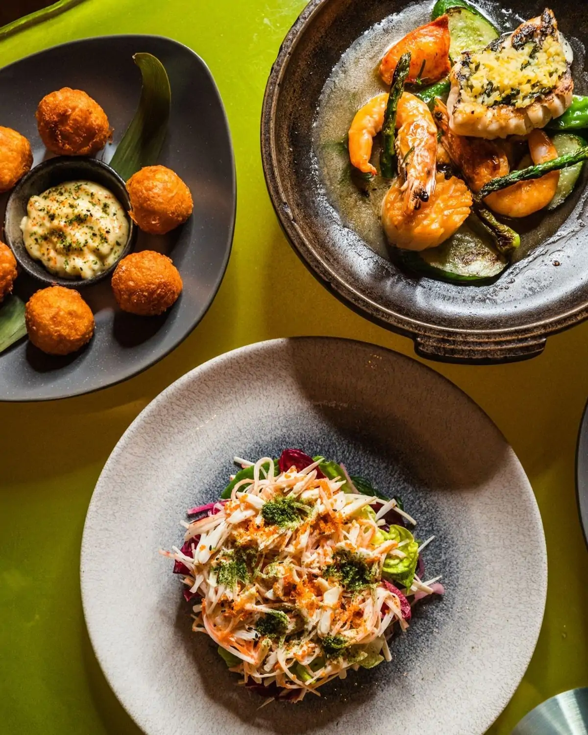 Overhead view of three appetizer plates on a green table, featuring fried balls with a creamy sauce, grilled shrimp and vegetables, and a salad with shredded toppings at Morimoto in Philadelphia on OpenTable.