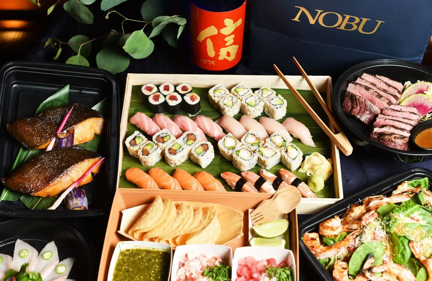 A premium takeout feast from Nobu Chicago, including their signature sushi rolls, fresh nigiri, flavorful cooked fish, unique tacos, and a bright shrimp and vegetable salad, accompanied by a bottle of sake, available to book on OpenTable.