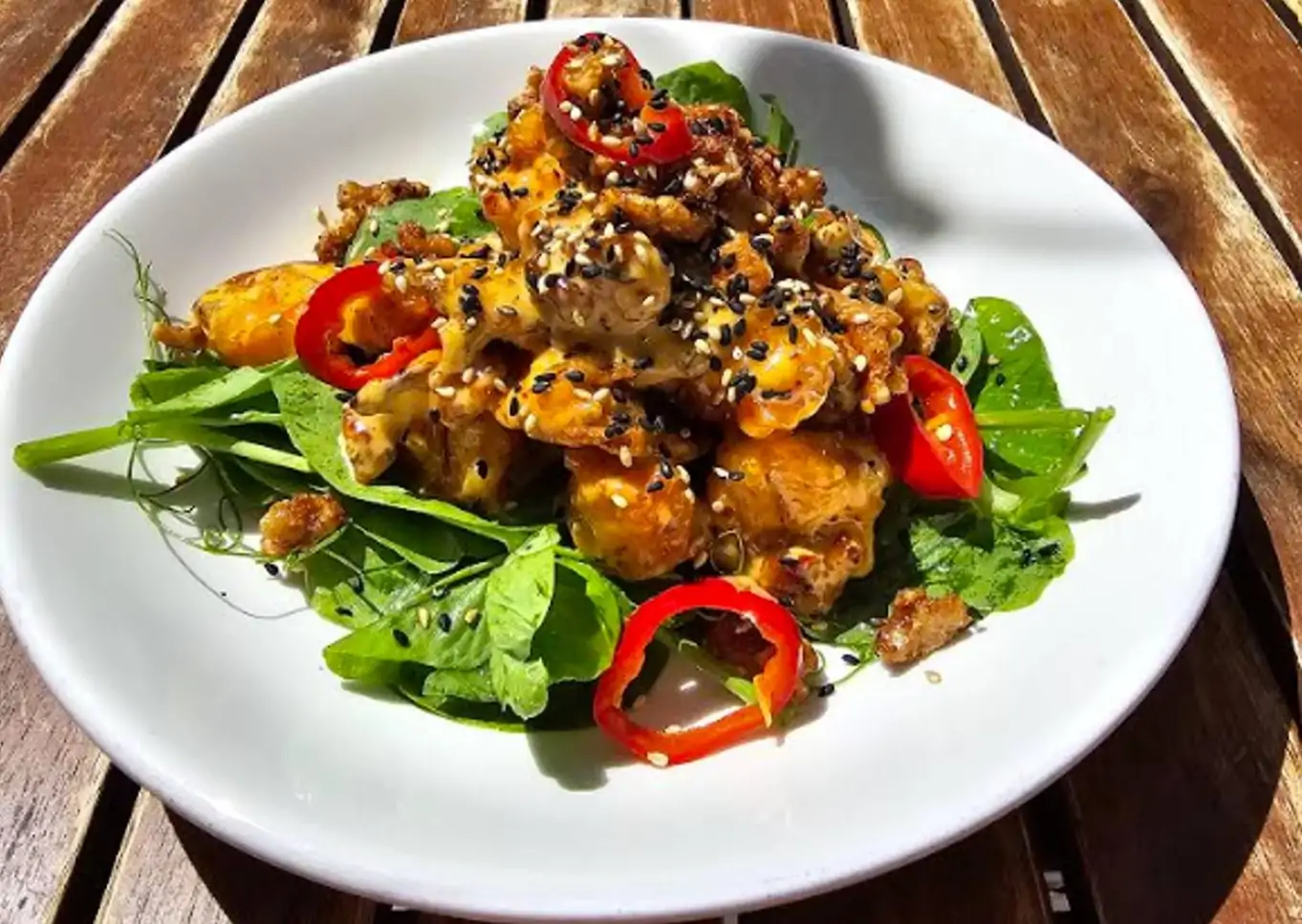 A close-up shows a white plate holding a mound of crispy shrimp with red chili slices in a glistening mayo sauce with black and white sesame seeds on a bed of greens at Ivan Ramen, one of the best solo dining NYC restaurants on OpenTable.