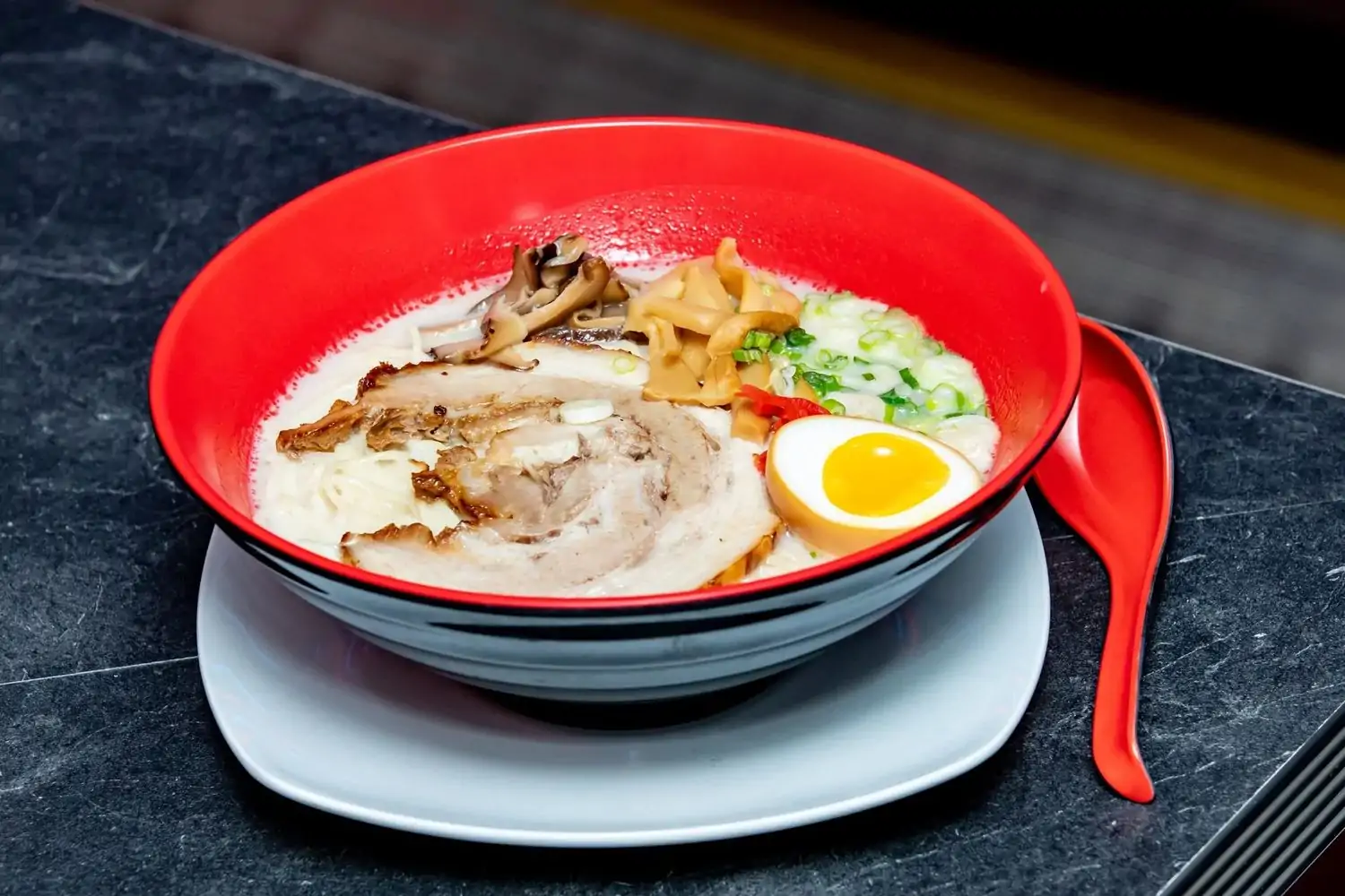 Close-up of a bowl of ramen with sliced pork belly, a soft-boiled egg, bamboo shoots, and chopped scallions in a creamy, opaque broth, served on a white plate with a red spoon at Chika Ramen Bar in Philadelphia. You can book on OpenTable.