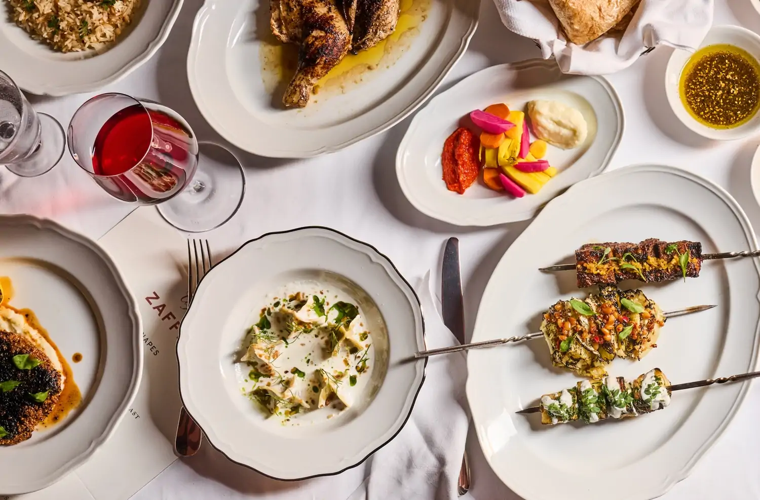 An aerial view shows a white cloth table topped with many white china plates holding dishes like kebabs, roasted chicken, rice, and colorful crudité with spreads and spiced dipping oils at Café Zaffri, a top solo dining NYC restaurant found on OpenTable.
