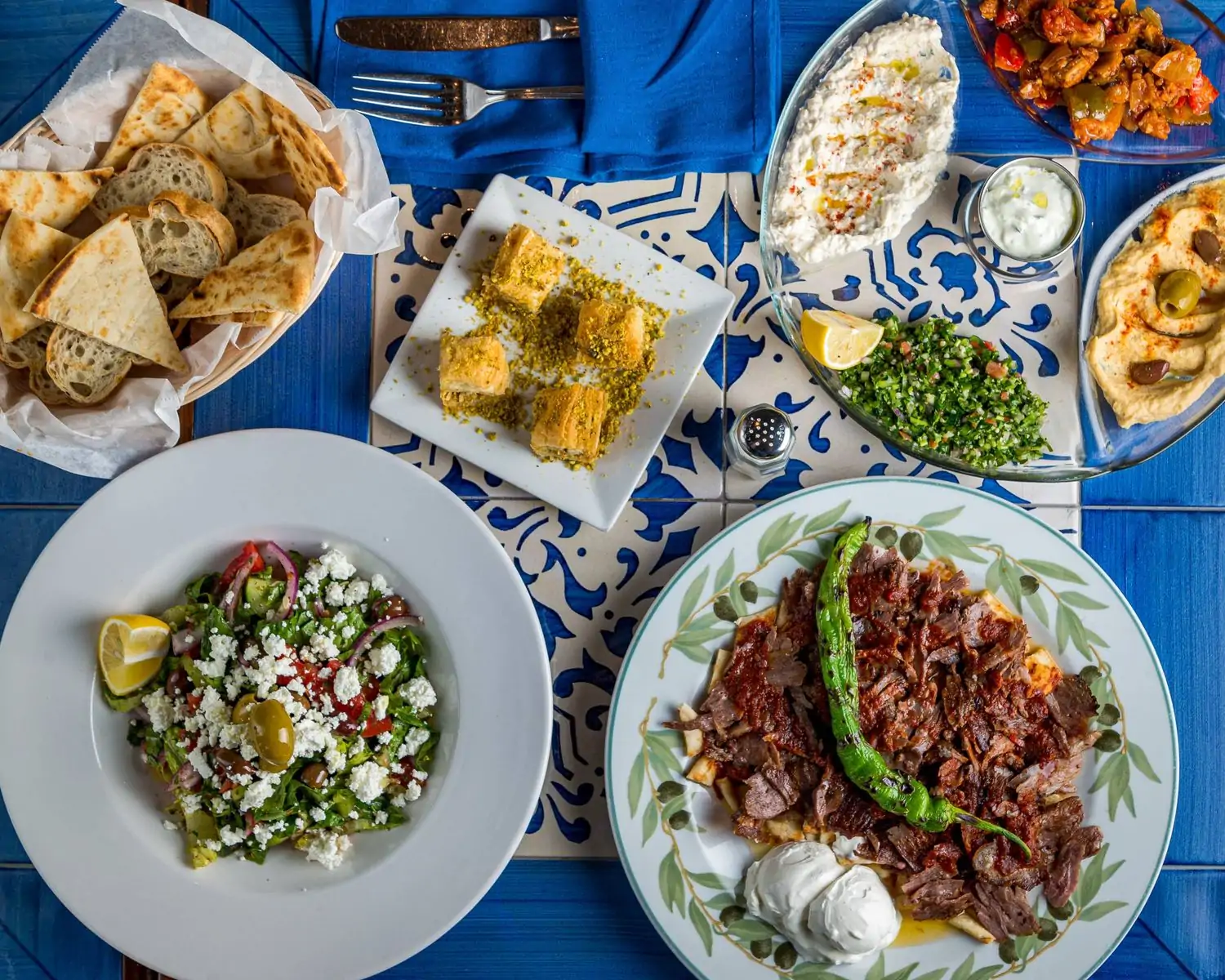 A blue and white tiled table set with Mediterranean mezze dishes, baklava, and pita at Kalamata Mediterranean Cuisine, in Miami. Book on OpenTable.