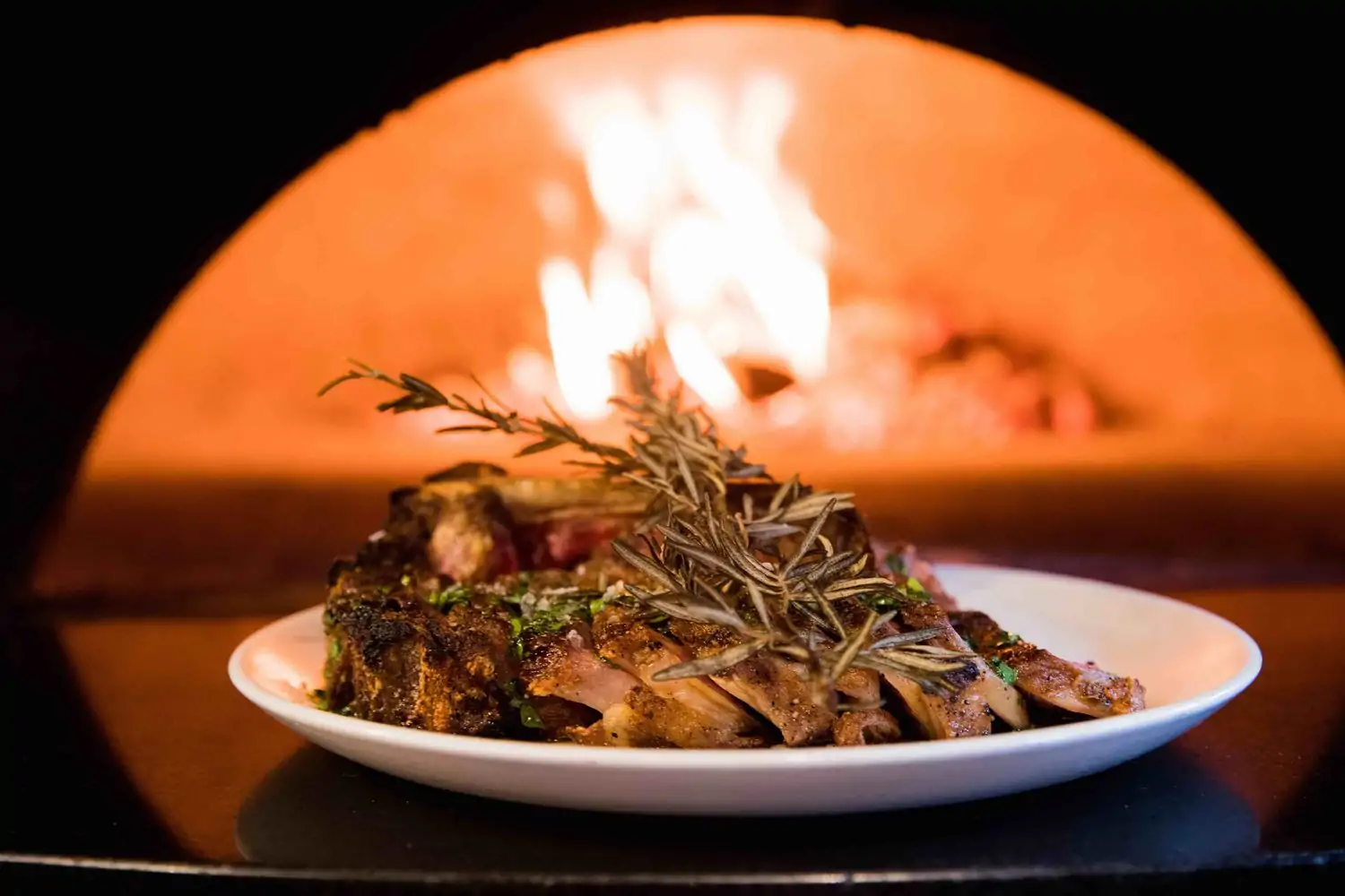 A white plate of grilled meat and rosemary in front of a wood-fired oven, available at Michael's Genuine Food & Drink, Miami. Book on OpenTable.