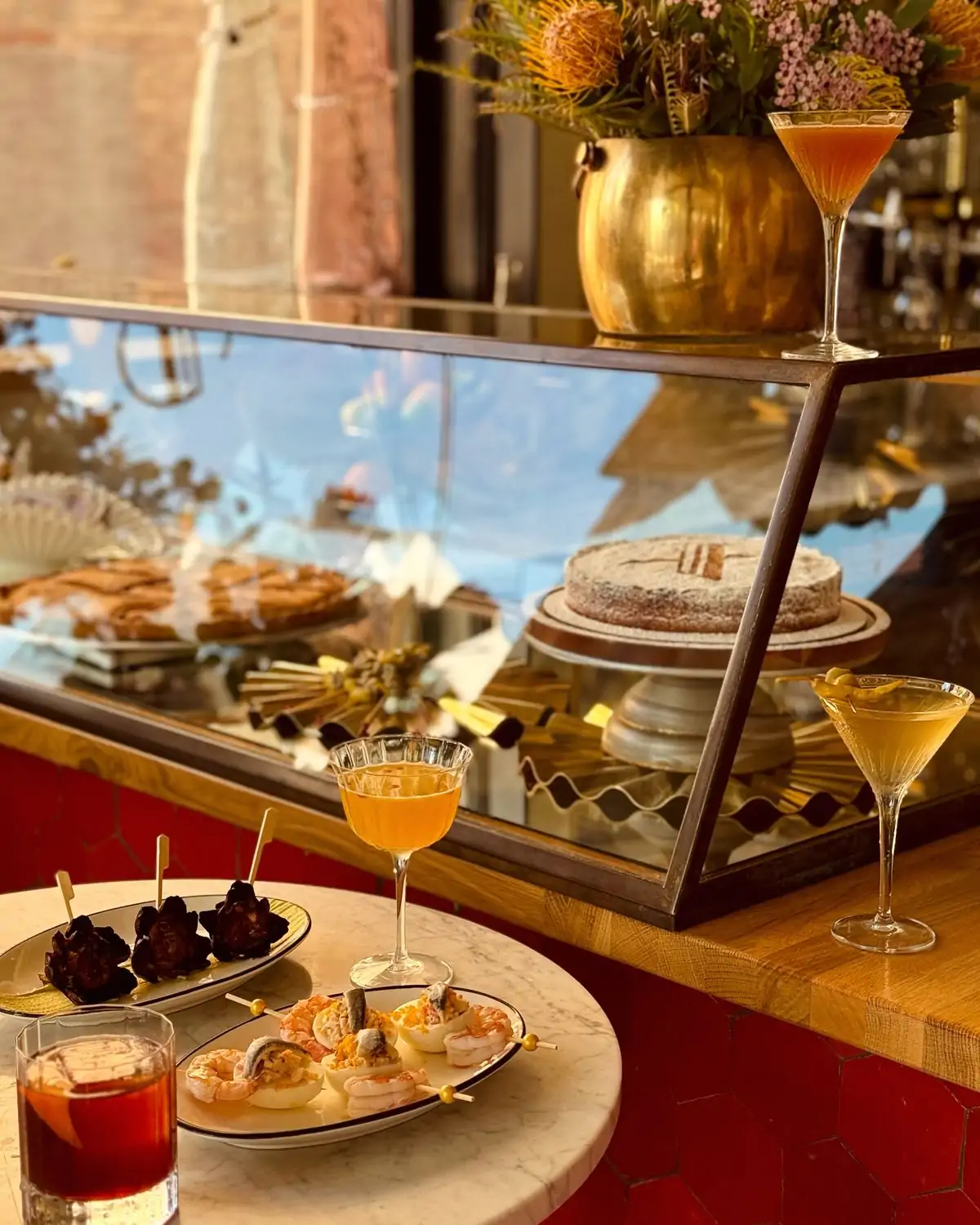 A table with skewered tapas and cocktails in front of a dessert case at Xuntos in Santa Monica. Book on OpenTable.