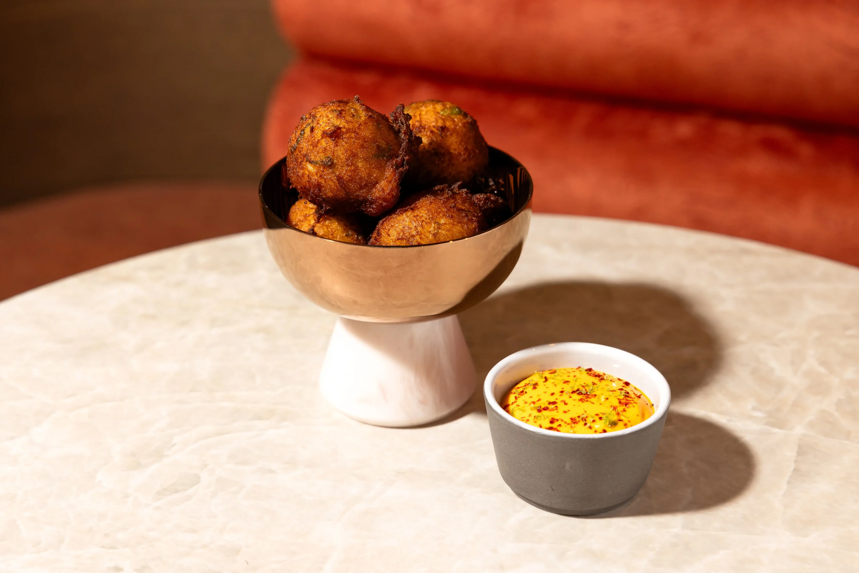A bowl of salted cod fritters with a side of sauce on a marble tabletop with a rust orange chair in the background at Haymarket in NYC on OpenTable.