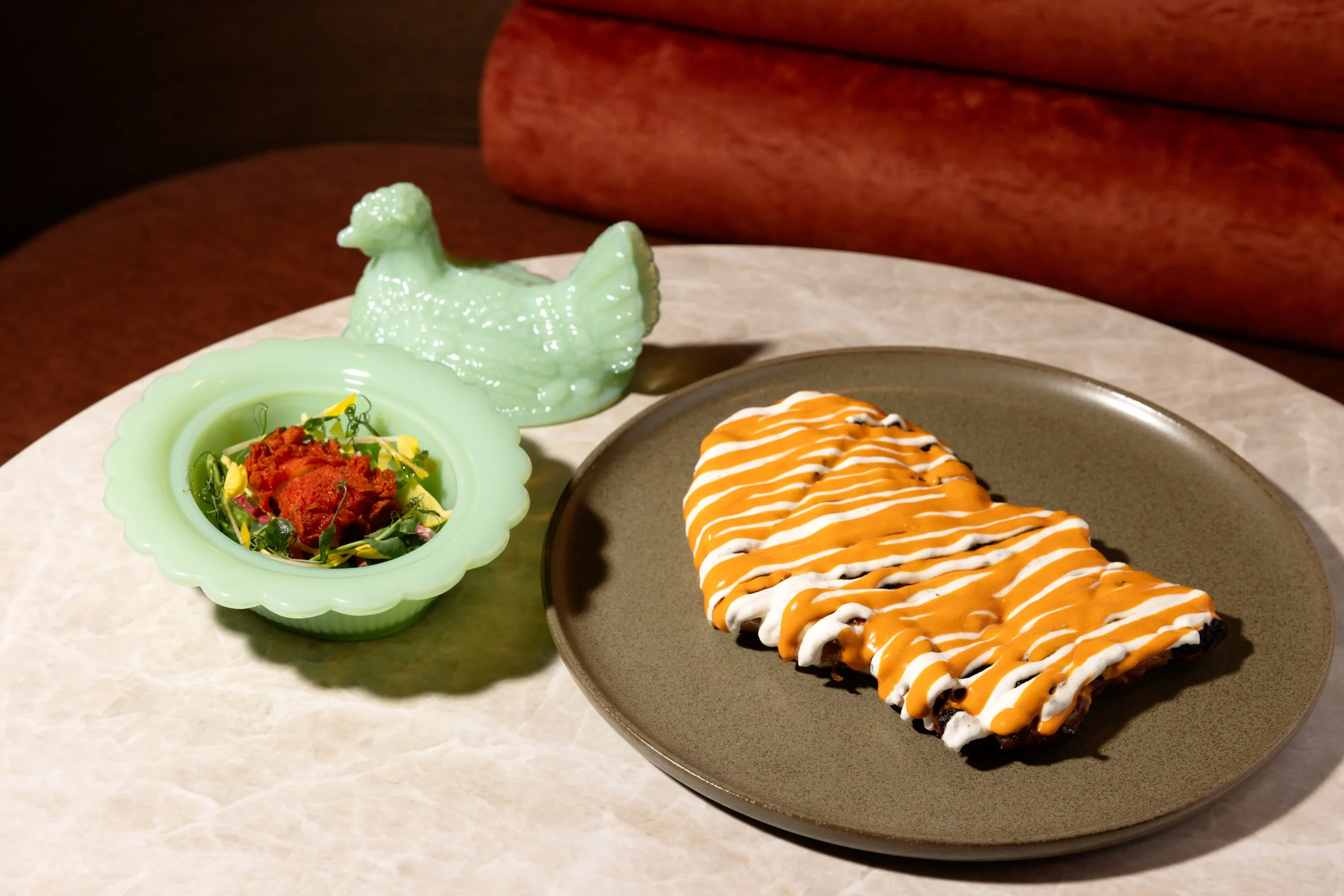 A brown ceramic plate of peri peri chicken with bright orange sauce drizzled and a mint green ceramic bird bowl dish with sauce and herbs at Haymarket in NYC. Book on OpenTable.