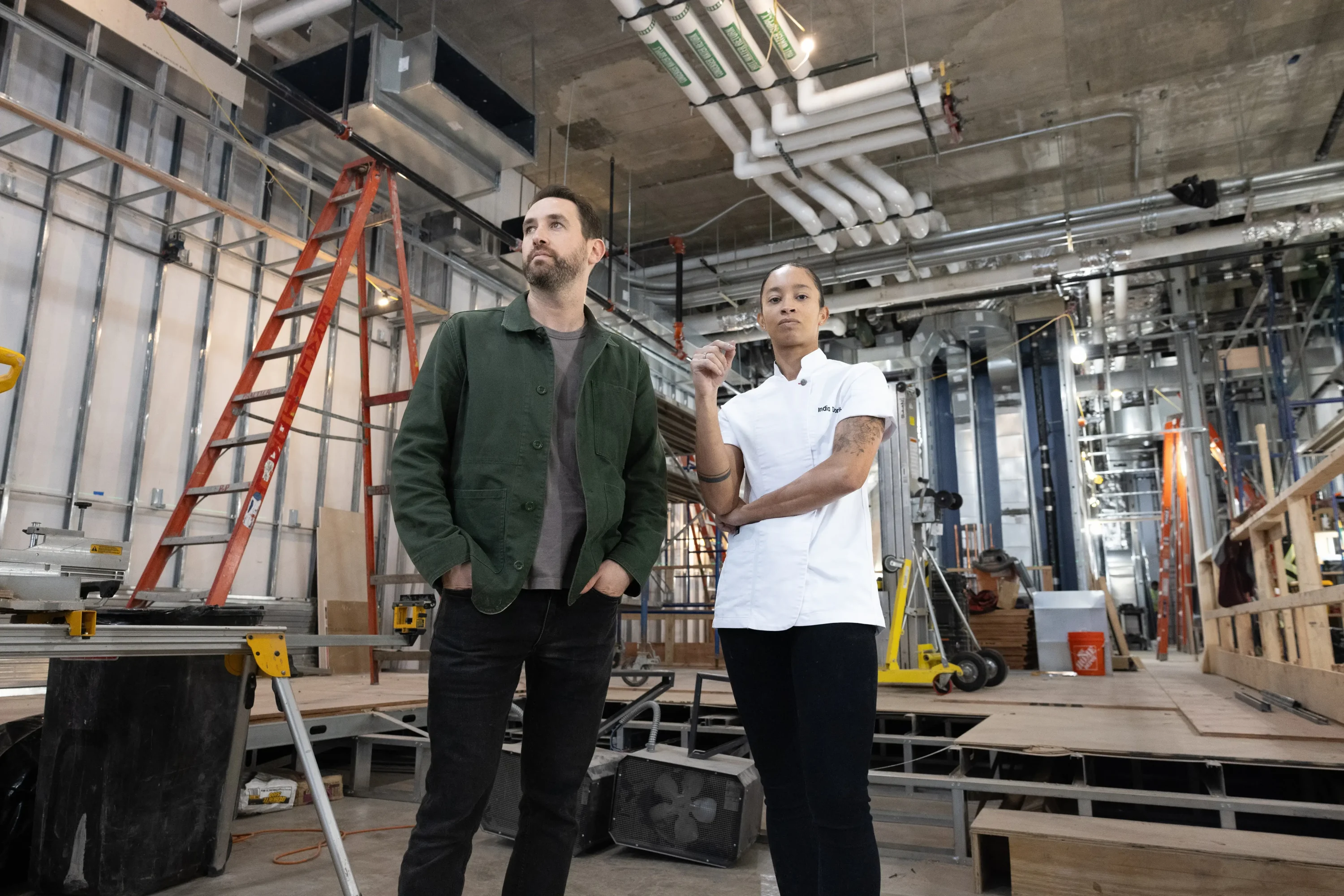 Alex Pfaffenbach, a man with a beard in a green jacket and black jeans and black and white sneakers, and India Doris, a woman in a white chef’s coat and black jeans with scaffolding and ladder behind them at Haymarket, available to book on OpenTable.