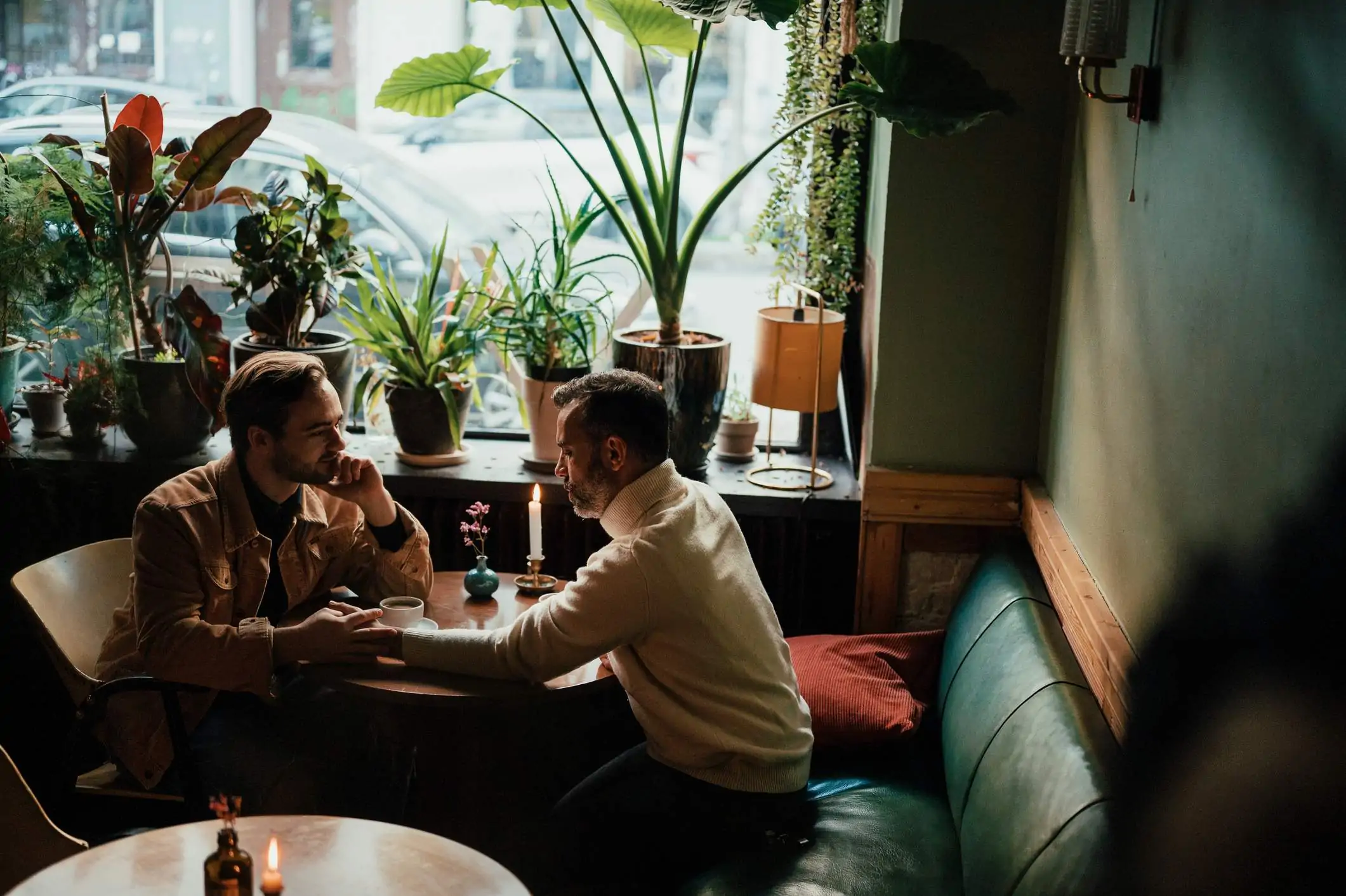 Image depicts two people sitting in a booth at a restaurant holding hands. One is wearing a turtleneck and the other has on a corduroy button up jacket. There are plants in the window next to them and they are drinking coffee.
