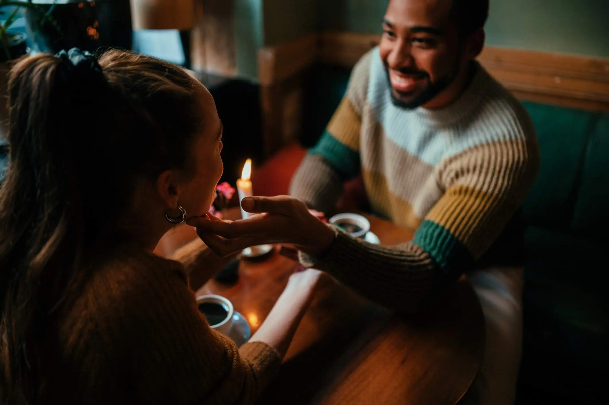 Image depicts two people sitting at a booth in a restaurant drinking coffee. One is wearing a striped turtleneck sweater and the other is wearing a brown sweater. The one in the striped sweater is reaching out to touch the face of the other person.
