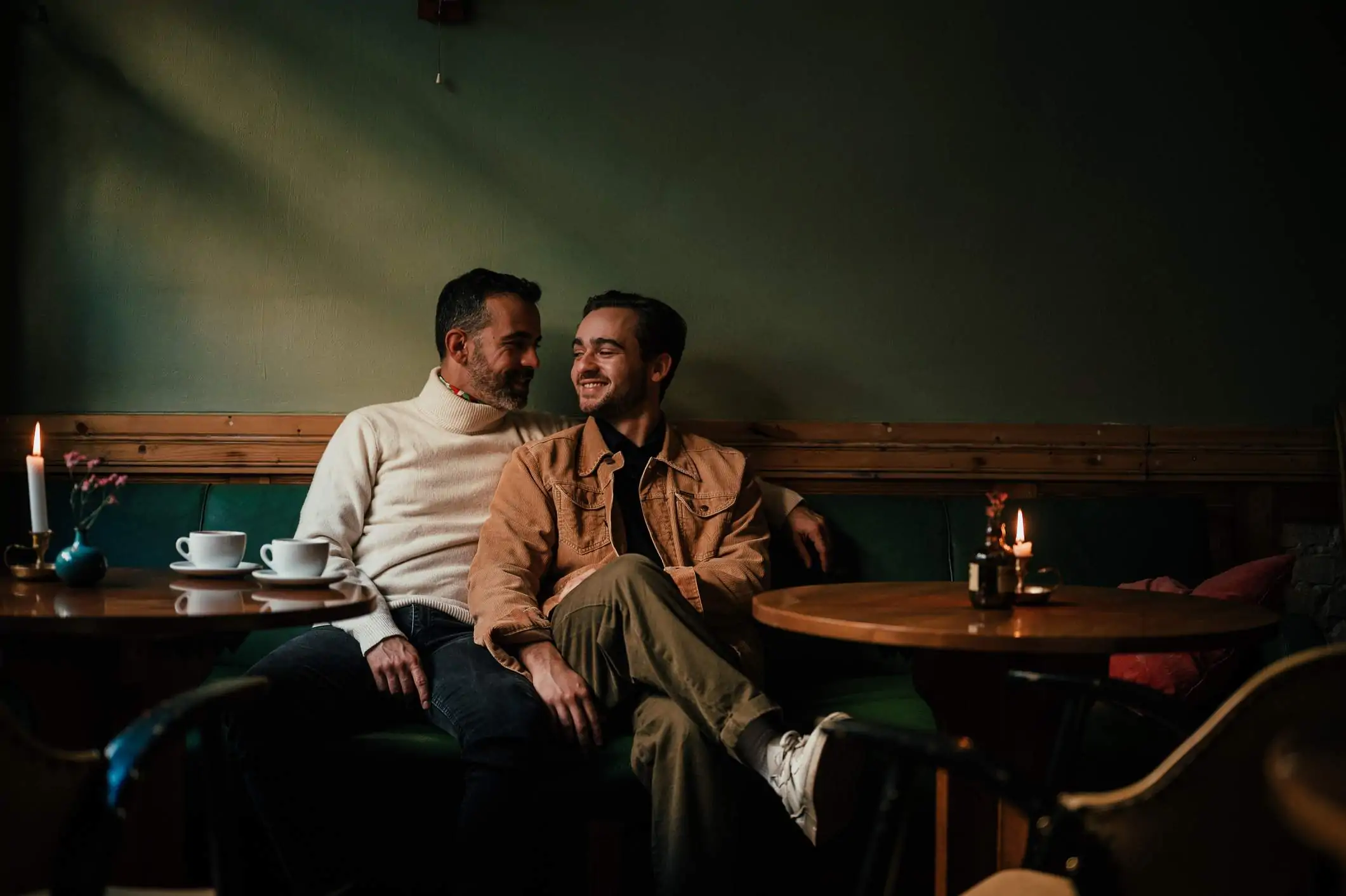 Image depicts two people cuddling in a booth at a restaurant. There are two coffee cups on a table in front of them.