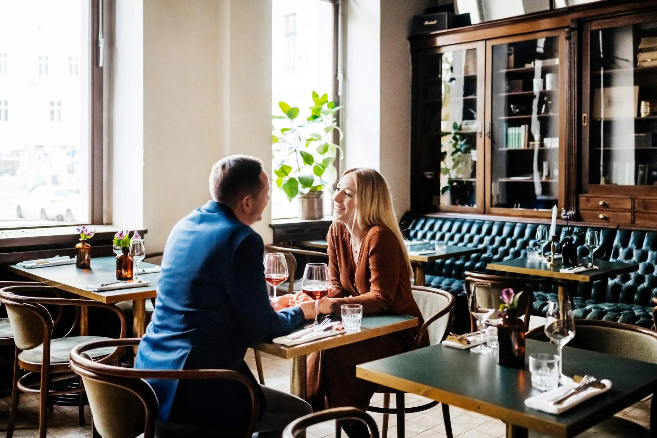 Image depicts a couple sitting together at a restaurant. They are holding hands, each drinking red wine, and smiling at each other.