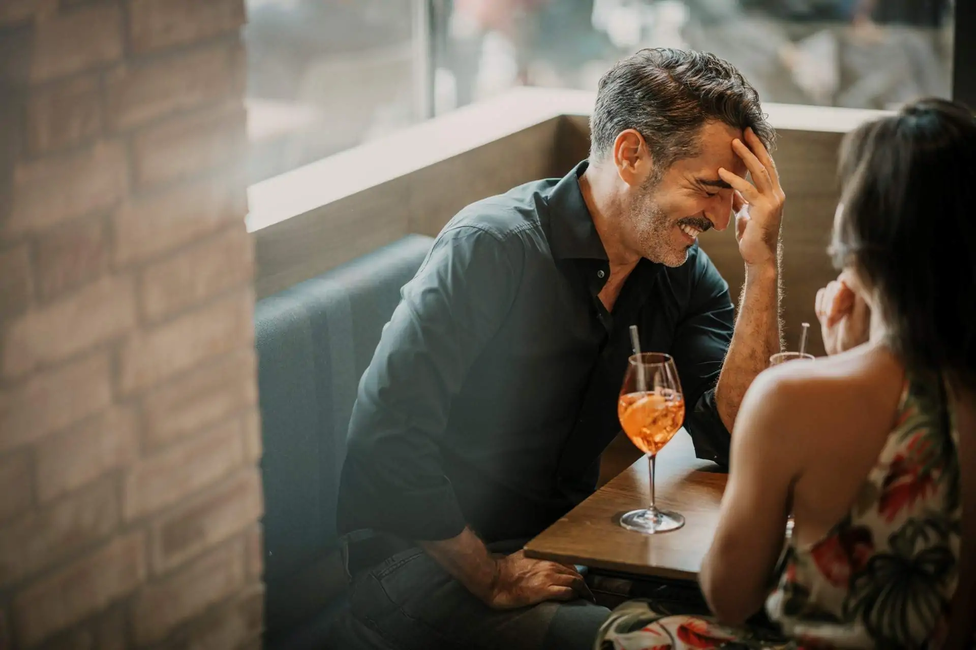 Image depicts two people sitting at a restaurant drinking orange drinks. One is laughing with their hand on their head.