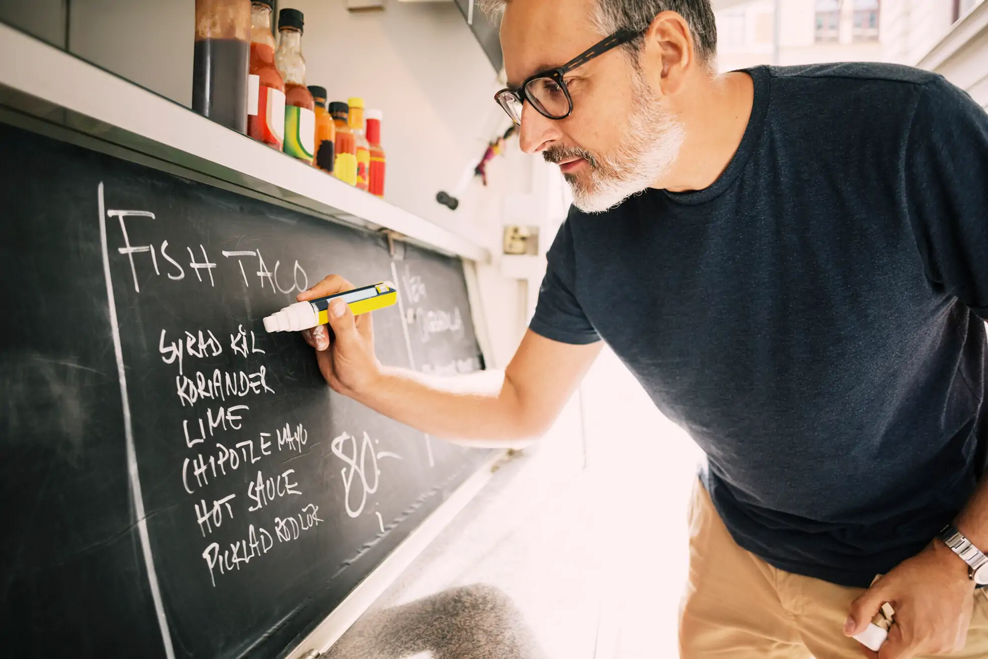 A chef writing the creative ingredients for a fish taco on a large chalkboard, representing the menu innovation and personality discussed in the article.