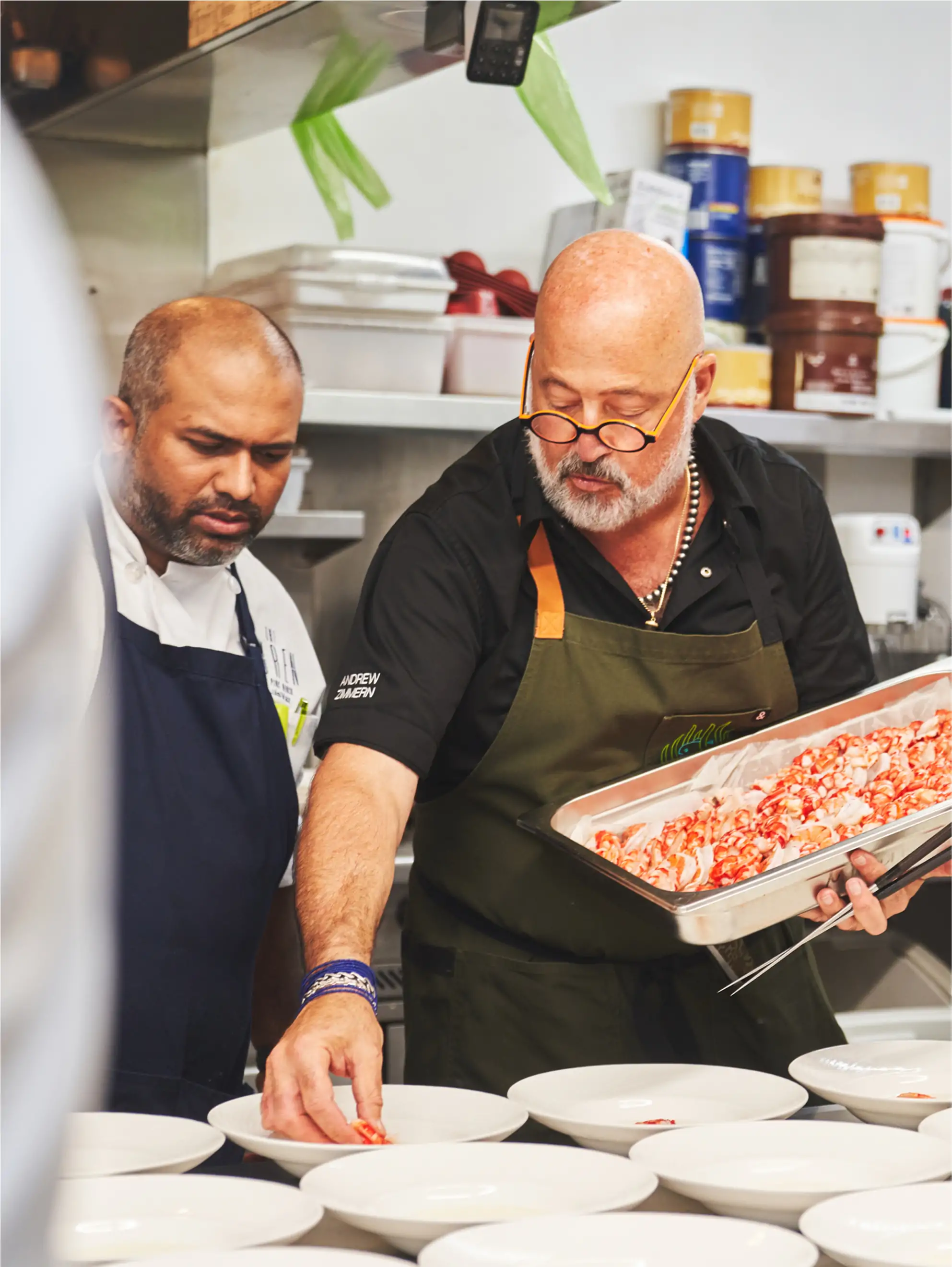 Author Andrew Zimmern in a professional kitchen, holding a tray of seafood and collaborating with another chef to plate dishes for service.