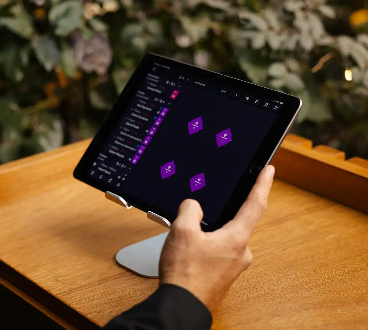 A person's hand holding an iPad on a stand, displaying a dark-themed restaurant management software interface with purple graphic elements.