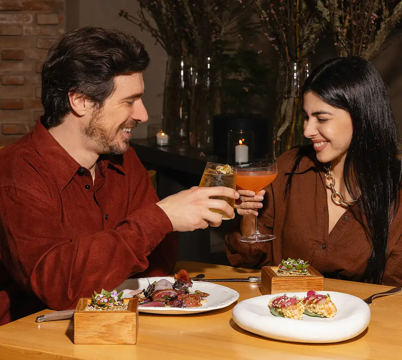 A smiling couple toasting with cocktails at a restaurant table, with modern, artistic dishes in front of them.