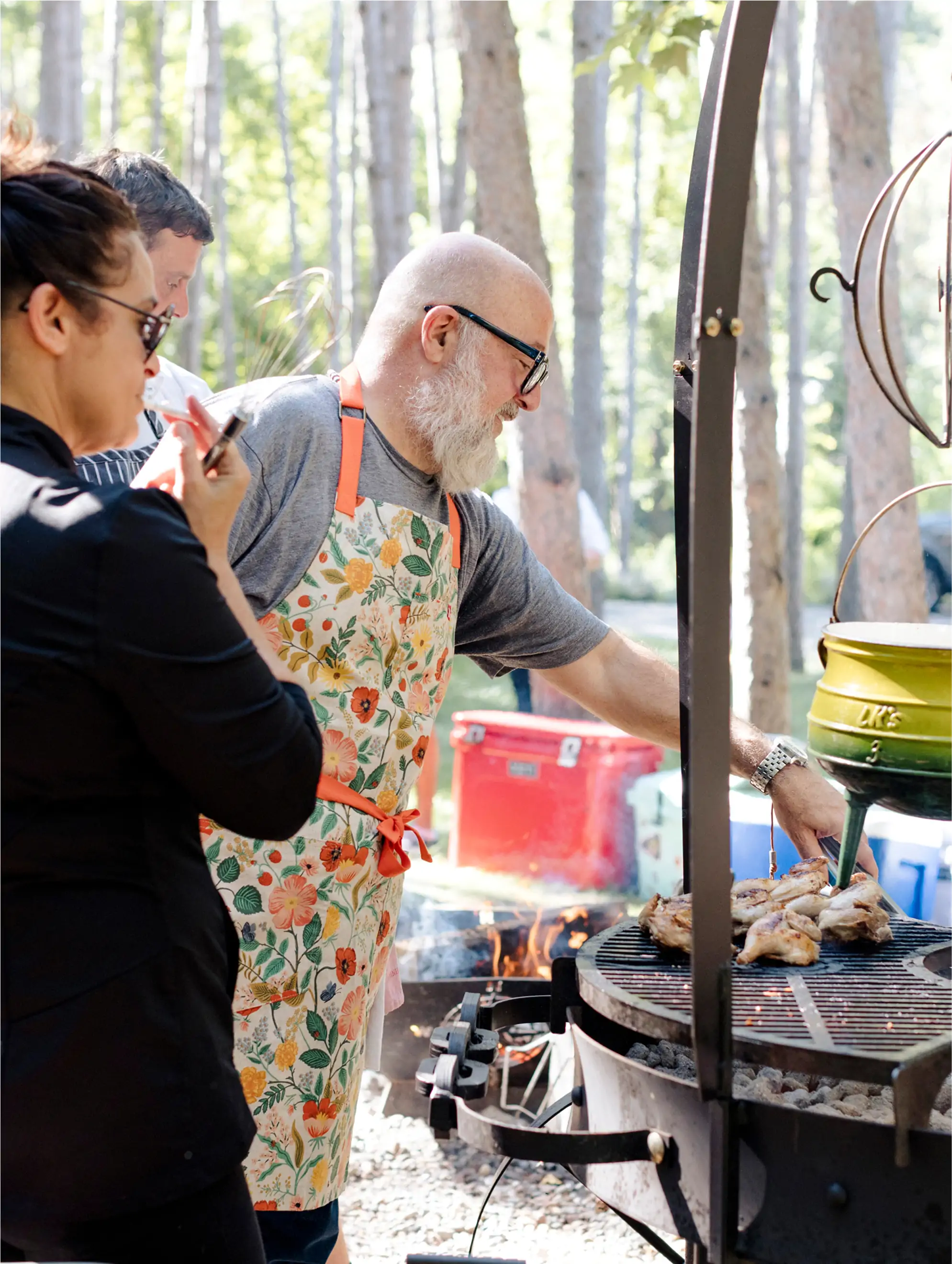 Author Andrew Zimmern, wearing a floral apron, grills chicken over an open fire at an outdoor event, demonstrating his passion for cooking in all its forms.