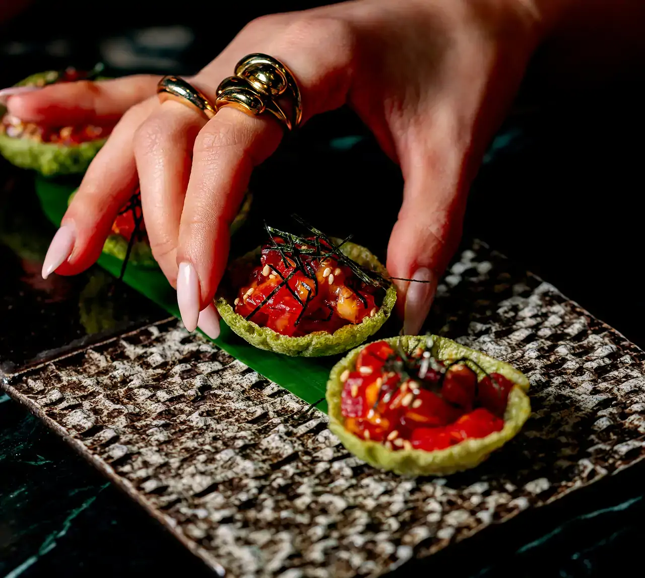 A hand with gold rings reaching for a green tartlet filled with red and white ingredients and garnished, presented on a patterned plate with another similar tartlet.